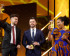 Nela Riehl, Member of the European Parliament for Volt, smiles joyfully while holding up her MEP Award on stage. She is wearing a vibrant blue and orange patterned dress. Two men in suits stand beside her, also holding awards and smiling, with a celebratory atmosphere and bright stage lighting in the background.