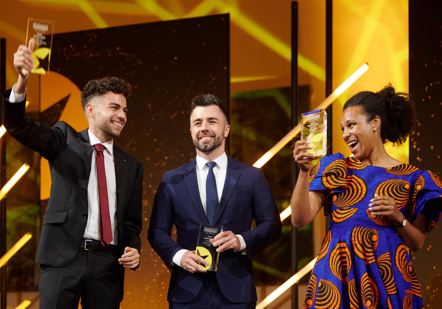 Nela Riehl, Member of the European Parliament for Volt, smiles joyfully while holding up her MEP Award on stage. She is wearing a vibrant blue and orange patterned dress. Two men in suits stand beside her, also holding awards and smiling, with a celebratory atmosphere and bright stage lighting in the background.