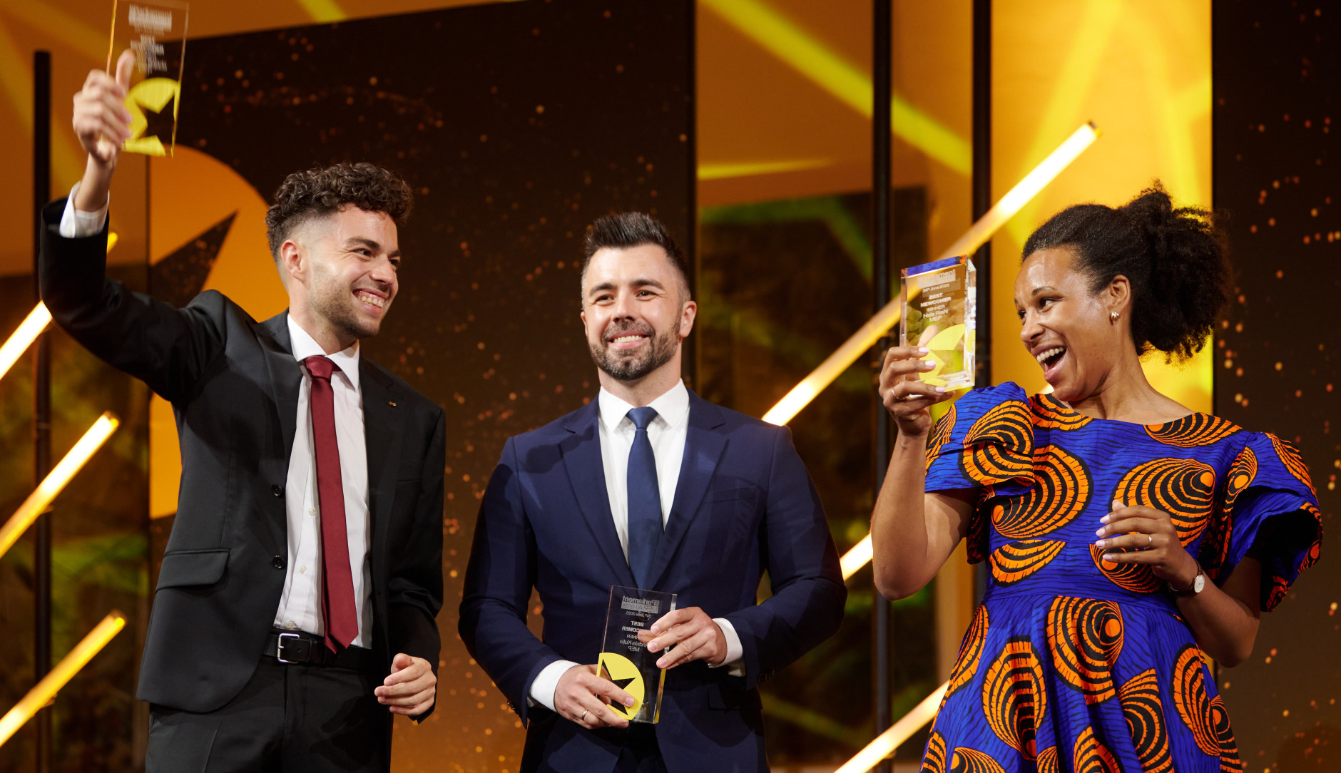 Nela Riehl, Member of the European Parliament for Volt, smiles joyfully while holding up her MEP Award on stage. She is wearing a vibrant blue and orange patterned dress. Two men in suits stand beside her, also holding awards and smiling, with a celebratory atmosphere and bright stage lighting in the background.