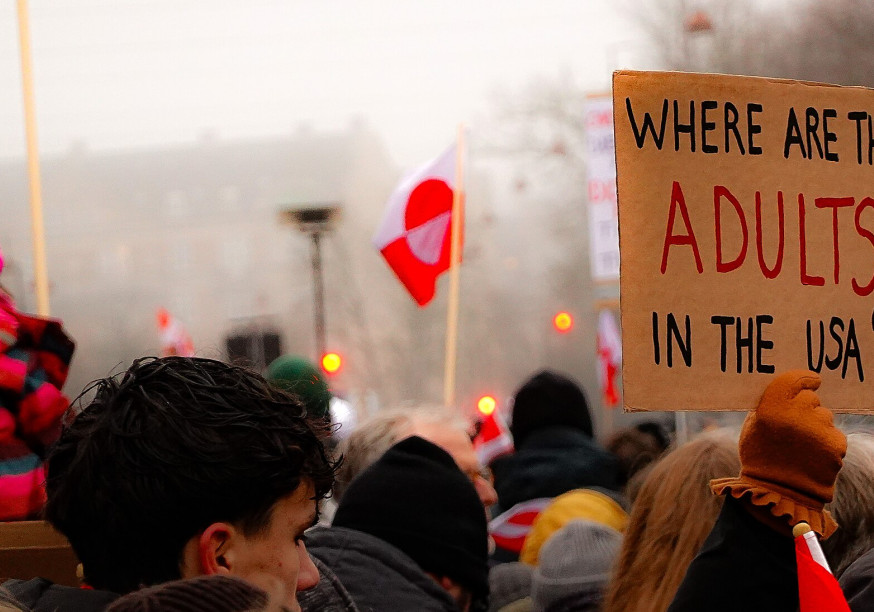 A close-up shot of a person from the side holding three small hand-held flags—the Greenlandic flag, the Danish Dannebrog, and the Faroese flag—against the backdrop of a large protest crowd. To the left, a white protest sign with red lettering says 
