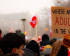 A close-up shot of a person from the side holding three small hand-held flags—the Greenlandic flag, the Danish Dannebrog, and the Faroese flag—against the backdrop of a large protest crowd. To the left, a white protest sign with red lettering says 