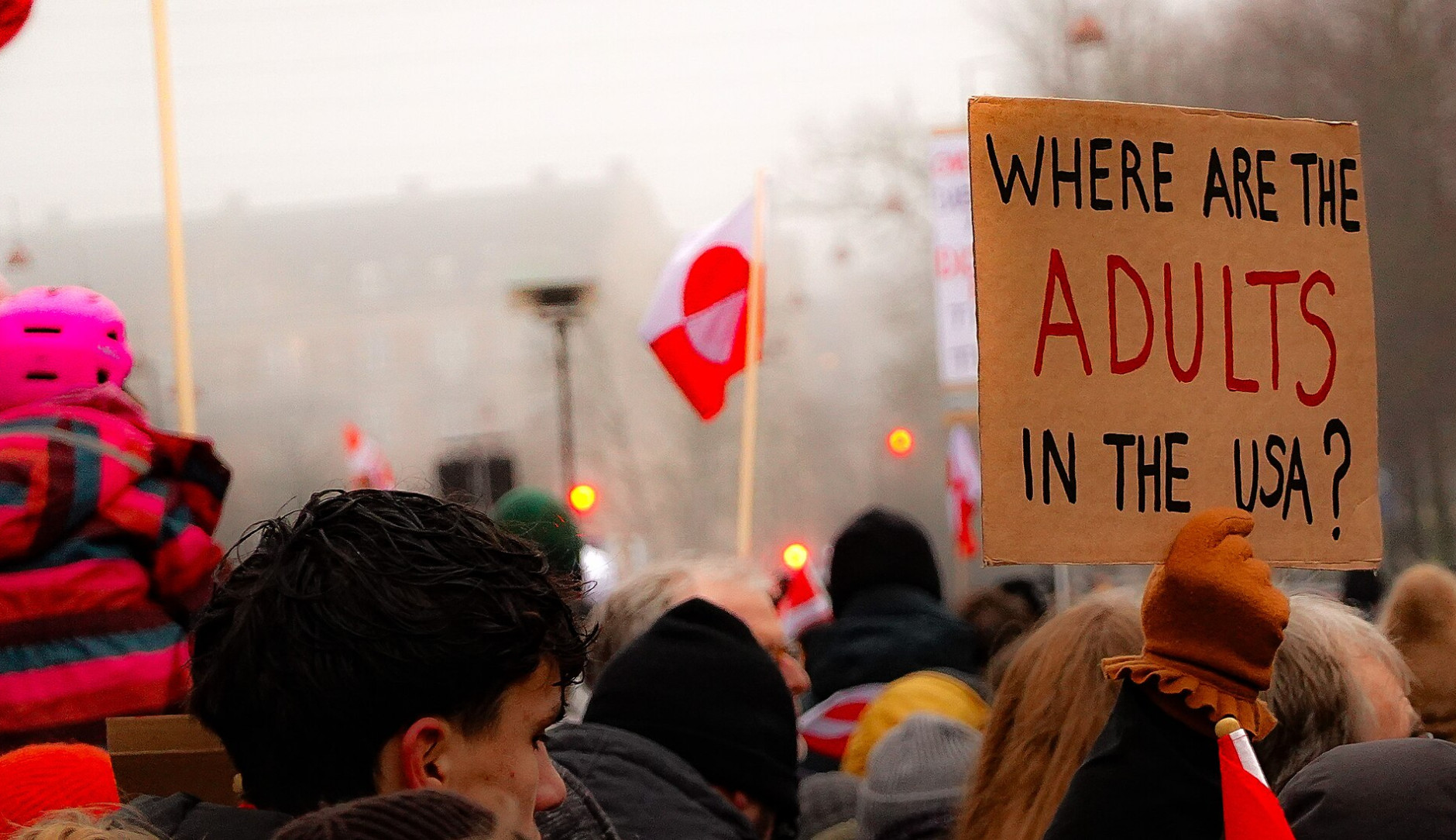 A close-up shot of a person from the side holding three small hand-held flags—the Greenlandic flag, the Danish Dannebrog, and the Faroese flag—against the backdrop of a large protest crowd. To the left, a white protest sign with red lettering says 