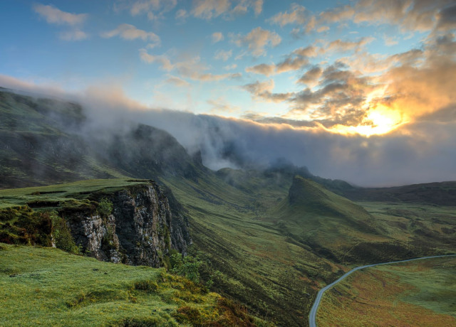 Natural landscape with a scenic route: the sky is blue but a bit cloudy with the sun peaking through. The green hills look luscious. No sign of humans except for a walkable road.