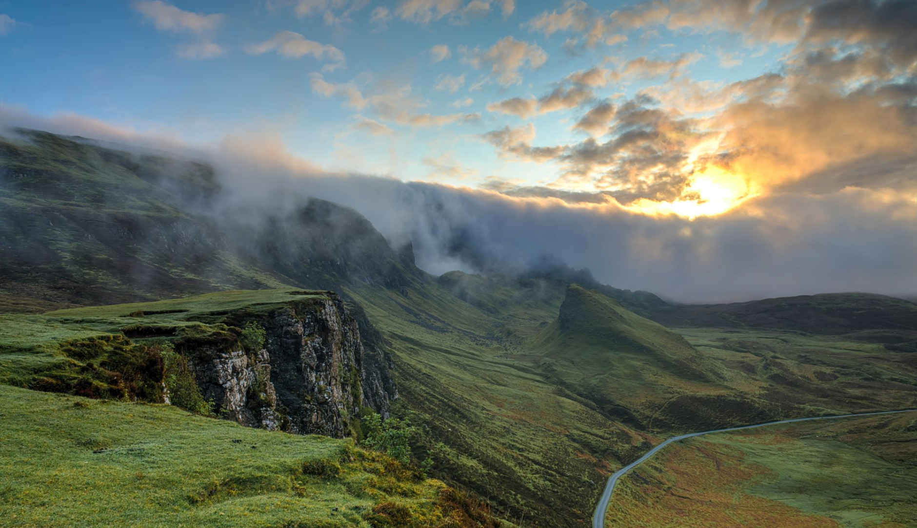 Natural landscape with a scenic route: the sky is blue but a bit cloudy with the sun peaking through. The green hills look luscious. No sign of humans except for a walkable road.