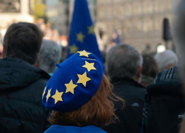 A woman with a blue beret decorated with the stars of the EU flag is seen in front of a EU flag.