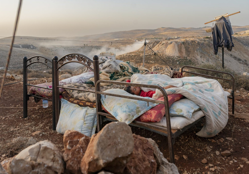 Source: https://mondoweiss.net/2024/11/photo-essay-the-silent-displacement-in-the-west-bank/#:~:text=As%20the%20world%20has%20focused,Bank%20following%20October%207%2C%202023. A photograph of two metal bed frames placed outdoors on a dry, rocky hillside. In the foreground bed, a young child is asleep under a thick, patterned blanket and pillows. The surrounding landscape is arid and mountainous, with a light haze or dust hanging in the valley behind them. Various items, including a water bottle and a jacket hanging from a wooden post, suggest a makeshift living space. The lighting is soft, indicating the early morning or late evening.
