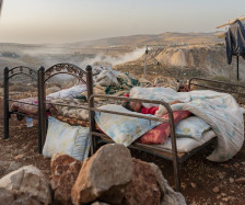 Source: https://mondoweiss.net/2024/11/photo-essay-the-silent-displacement-in-the-west-bank/#:~:text=As%20the%20world%20has%20focused,Bank%20following%20October%207%2C%202023. A photograph of two metal bed frames placed outdoors on a dry, rocky hillside. In the foreground bed, a young child is asleep under a thick, patterned blanket and pillows. The surrounding landscape is arid and mountainous, with a light haze or dust hanging in the valley behind them. Various items, including a water bottle and a jacket hanging from a wooden post, suggest a makeshift living space. The lighting is soft, indicating the early morning or late evening.
